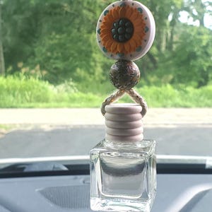 May include: A clear, square glass bottle with a wooden top, suspended by a rope. Above the bottle is a decorative sunflower bead and a patterned bead. The car air freshener hangs in front of a blurred green background.