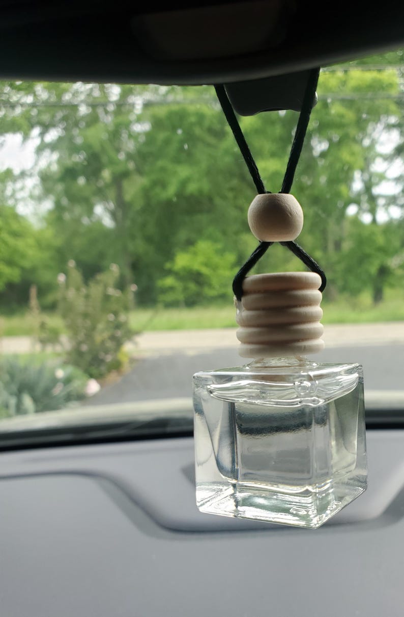 K&ouml;nnte beinhalten: Ein klarer, quadratischer Auto-Lufterfrischer aus Glas mit einem Holzaufsatz und einer Holzperle, die an einer schwarzen Schnur h&auml;ngt. Der Lufterfrischer ist mit einer klaren Fl&uuml;ssigkeit gef&uuml;llt. Der Hintergrund ist eine verschwommene Ansicht von gr&uuml;nen B&auml;umen.