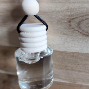 K&ouml;nnte beinhalten: Eine klare Glas-Lufterfrischerflasche f&uuml;r Autos mit einem hellen holzfarbenen Deckel und einer schwarzen Schnur zum Aufh&auml;ngen. Die Flasche ist mit einer klaren Fl&uuml;ssigkeit gef&uuml;llt. Der Hintergrund ist ein hellbraunes Holzmaserung.