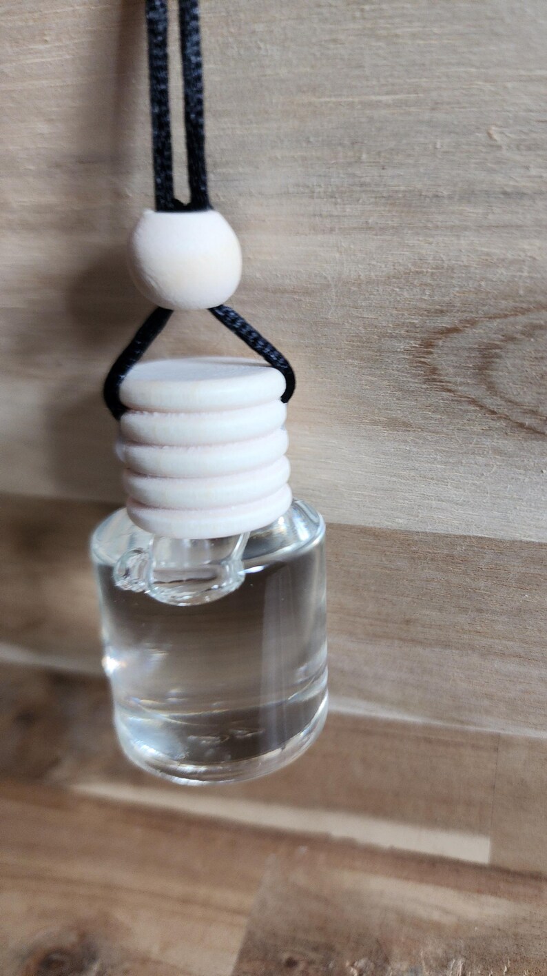 K&ouml;nnte beinhalten: Eine klare Glas-Lufterfrischerflasche f&uuml;r Autos mit einem hellen holzfarbenen Deckel und einer schwarzen Schnur zum Aufh&auml;ngen. Die Flasche ist mit einer klaren Fl&uuml;ssigkeit gef&uuml;llt. Der Hintergrund ist ein hellbraunes Holzmaserung.