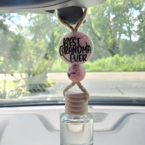 May include: An air freshener hanging from a car's rearview mirror. The top bead is white with the text "BEST GRANDMA EVER" and a pink heart. Below is a pink bead, and a small glass bottle with a wooden cap.