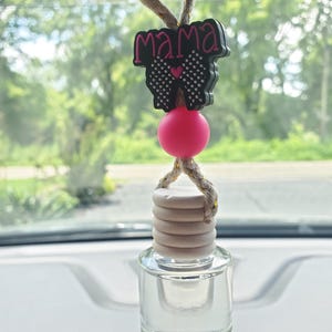 May include: A car air freshener with a clear glass bottle and a wooden cap. The top features a black charm with the word "mama" in pink, a bow, and a pink bead. The freshener hangs from a beige rope.