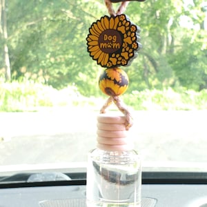 May include: A car air freshener with a clear glass bottle and a wooden cap. The air freshener is suspended by a string and features a sunflower charm with the words "Dog Mom" and paw prints. A decorative bead is below the charm.