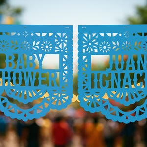 May include: Two blue paper banners with intricate cut-out designs and scalloped edges. The left banner reads "BABY SHOWER" and the right banner reads "JUAN IGNACIO". The banners are strung together, perfect for a baby shower celebration.