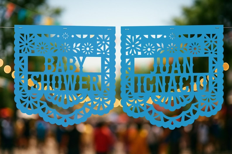 May include: Two blue paper banners with intricate cut-out designs and scalloped edges. The left banner reads "BABY SHOWER" and the right banner reads "JUAN IGNACIO". The banners are strung together, perfect for a baby shower celebration.