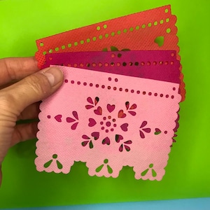 May include: A stack of colorful, decorative paper cutouts in shades of red, pink, and magenta. The cutouts feature intricate designs of hearts, dots, and scalloped edges. The paper is held against a bright green background.