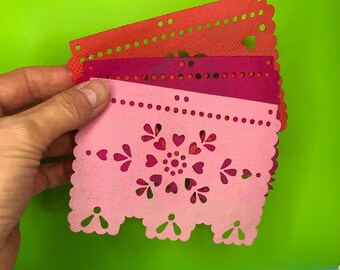 May include: A stack of colorful, decorative paper cutouts in shades of red, pink, and magenta. The cutouts feature intricate designs of hearts, dots, and scalloped edges. The paper is held against a bright green background.
