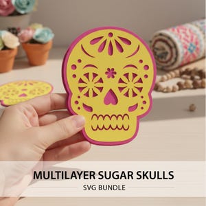 May include: A layered sugar skull design in yellow and pink, held by a hand. The skull features intricate cut-out details and a decorative floral pattern. The text "MULTILAYER SUGAR SKULLS SVG BUNDLE" is displayed at the bottom.