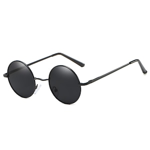 Puede incluir: Gafas de sol redondas negras con una fina montura de metal negro. Las lentes oscuras tienen forma circular y las patillas se extienden hacia los lados. Las gafas tienen un estilo retro.