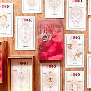 Peut inclure: Un jeu de cartes de tarot avec un design rose et or. Les cartes pr&eacute;sentent une vari&eacute;t&eacute; de symboles et d'images, notamment des lunes, des &eacute;toiles et des formes g&eacute;om&eacute;triques. Les cartes sont dispos&eacute;es en cercle autour d'un livre intitul&eacute; "Destiny Deck: The Art of Tarot".
