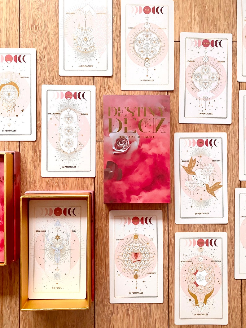 Peut inclure: Un jeu de cartes de tarot avec un design rose et or. Les cartes pr&eacute;sentent une vari&eacute;t&eacute; de symboles et d'images, notamment des lunes, des &eacute;toiles et des formes g&eacute;om&eacute;triques. Les cartes sont dispos&eacute;es en cercle autour d'un livre intitul&eacute; "Destiny Deck: The Art of Tarot".