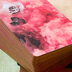 Peut inclure: Un jeu de cartes de tarot rose et or avec une rose blanche et un cristal sur la carte du dessus. Les cartes ont un fond aquarelle rose et blanc avec une subtile brillance.