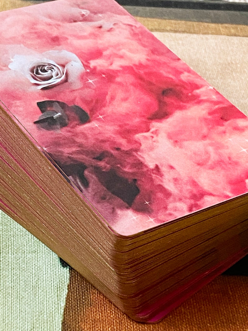 Peut inclure: Un jeu de cartes de tarot rose et or avec une rose blanche et un cristal sur la carte du dessus. Les cartes ont un fond aquarelle rose et blanc avec une subtile brillance.