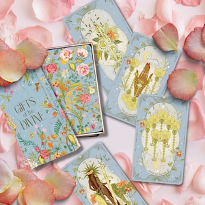 May include: A blue box with the text "Gifts of the Divine" on the front. The box contains a deck of tarot cards with illustrations of flowers, hands, and wands. The cards are on a background of pink rose petals.