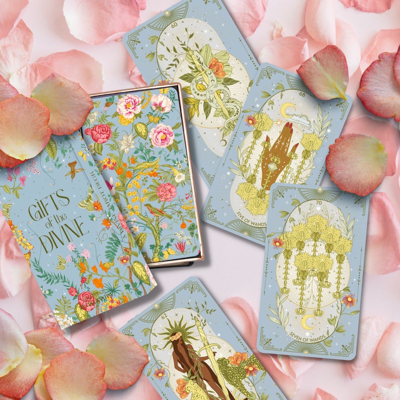 May include: A blue box with the text "Gifts of the Divine" on the front. The box contains a deck of tarot cards with illustrations of flowers, hands, and wands. The cards are on a background of pink rose petals.