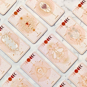 Peut inclure: Un jeu de cartes de tarot avec un design rose et or. Chaque carte pr&eacute;sente une illustration diff&eacute;rente et le nom de la carte. Les cartes sont dispos&eacute;es en cercle sur une surface en marbre blanc.