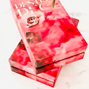 Peut inclure: Trois bo&icirc;tes empil&eacute;es d'un jeu de tarot appel&eacute; "Destiny Deck: The Art of Tarot". Les bo&icirc;tes sont roses avec un design aquarelle de nuages et une rose.