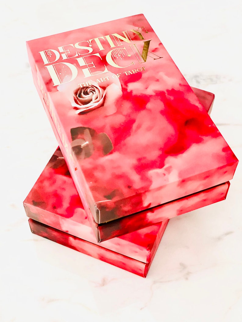 Peut inclure: Trois bo&icirc;tes empil&eacute;es d'un jeu de tarot appel&eacute; "Destiny Deck: The Art of Tarot". Les bo&icirc;tes sont roses avec un design aquarelle de nuages et une rose.