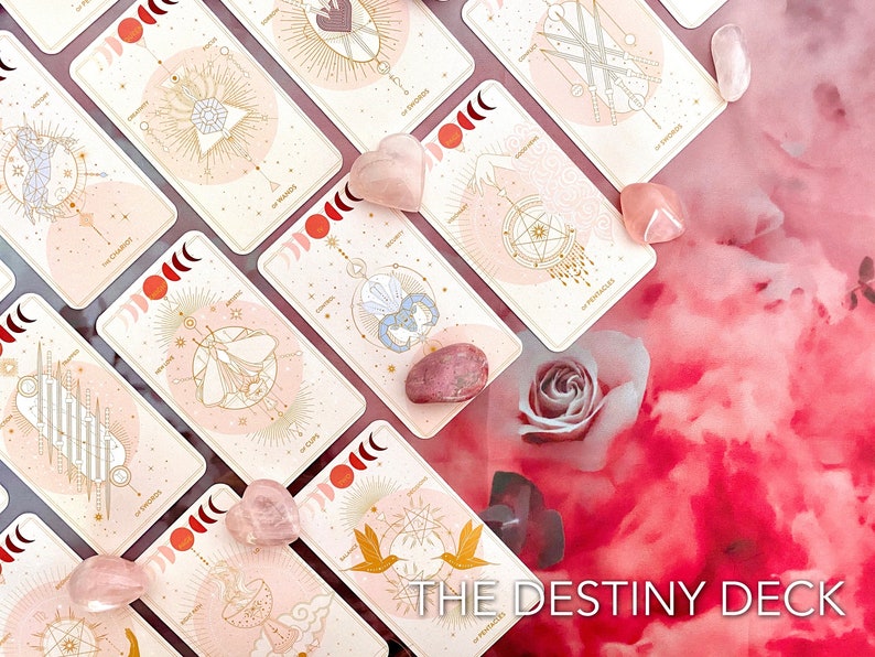 Peut inclure: Un jeu de cartes de tarot avec un design rose et or. Les cartes sont dispos&eacute;es sur un fond rose et blanc avec des cristaux de quartz rose et une seule rose. Le texte "THE DESTINY DECK" est visible en bas de l'image.