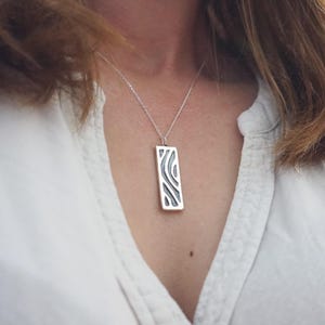 Puede incluir: Un collar con colgante de plata con un colgante rectangular. El colgante presenta un diseño oscuro y abstracto dentro de un marco plateado. El collar se lleva sobre una camisa blanca.