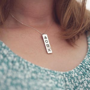 Peut inclure: Un collier pendentif argenté avec un pendentif rectangulaire. Le pendentif présente un triangle, un cercle, un carré et un symbole X. Le collier est porté par une personne avec un haut floral vert et blanc.