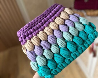 May include: A handmade, colorful clutch with a textured, woven design. The clutch features a gradient of colors, including purple, tan, light purple, teal, and turquoise. The top of the clutch is a solid purple, while the bottom transitions to turquoise.