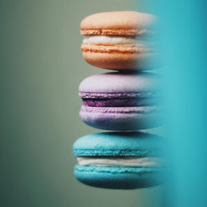 Peut inclure: Une pile de trois macarons colorés. Le macaron du haut est orange, celui du milieu est violet et celui du bas est turquoise. Chaque macaron a une garniture blanche. Le fond est vert.