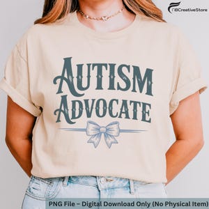 Puede incluir: Camiseta color crema con las palabras "AUTISM ADVOCATE" en una fuente estilizada de color verde azulado oscuro. Un lazo azul claro está centrado debajo del texto. La camiseta la lleva una persona con las mangas dobladas.