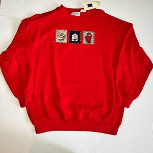 May include: A bright red sweatshirt with three embroidered patches. The patches feature a sprig of holly, a birdhouse, and a red mitten. The sweatshirt has a crew neck and long sleeves. A tag is attached to the neckline.