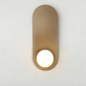 May include: A modern wall sconce with a gold-toned, elongated oval backplate. A round, glowing light source is positioned at the bottom. The sconce is mounted on a textured white wall, providing a minimalist aesthetic.
