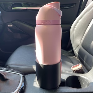 May include: A light pink Owala water bottle with a matching pink lid and a dark pink band featuring the brand name. The bottle is placed inside a black cylindrical cup holder adapter, which is positioned within a vehicle center console.
