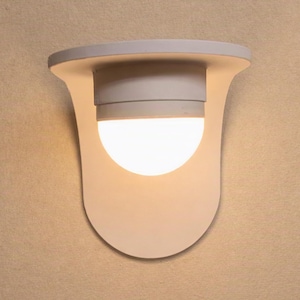 May include: A white wall-mounted light fitting featuring a curved, shield-shaped backplate and a central cylindrical housing that supports a glowing, semi-spherical glass bulb. The design is smooth and uniform, emitting a soft, warm light.