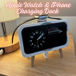 Può includere: Una base di ricarica bianca per Apple Watch e iPhone. L'iPhone mostra un quadrante dell'orologio. La base ha un design minimalista con piccole gambe in legno e spazio per l'orologio.