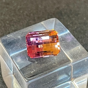 Puede incluir: Una gema de corte rectangular con un degradado de colores, que va del rosa al naranja. La gema está colocada sobre una superficie clara y reflectante, mostrando sus facetas y estructura interna. Los tonos vibrantes crean un efecto visual llamativo.