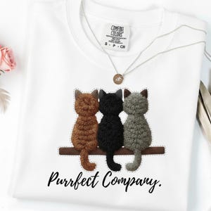 Könnte beinhalten: Weißes Sweatshirt mit einer Grafik von drei Katzen in Braun, Schwarz und Grau, die auf einem braunen Holzbrett sitzen. Der Text "Purrfect Company" steht darunter. Eine silberne Halskette mit Anhänger befindet sich auf dem Shirt.