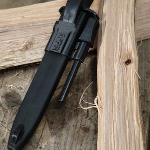 Puede incluir: Un cuchillo negro con funda y un iniciador de fuego. El cuchillo está colocado en diagonal sobre dos trozos de madera clara. La funda tiene las letras "APR" impresas. El cuchillo está diseñado para uso en exteriores.