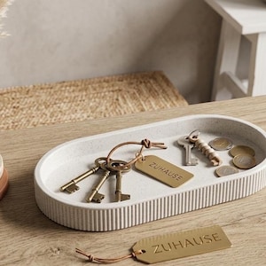 Op de afbeelding: Een decoratief dienblad met sleutels, munten en een label met de tekst "ZUHAUSE". Een kleine zwarte vaas met een gedroogde plant en een stapel onderzetters staan ook op het houten oppervlak. Een bril en een klein leren notitieboekje zijn ook zichtbaar.