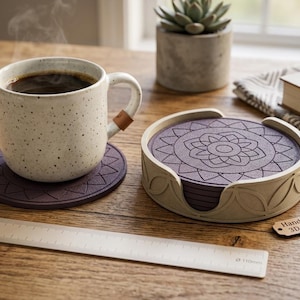 May include: A speckled ceramic mug with a leather accent on the handle sits on a purple coaster. A set of matching coasters with a floral design is in a wooden holder. A ruler and a small succulent plant are also visible.