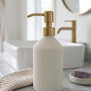 Puede incluir: Dispensador de jabón de cerámica color crema con detalles de estrías verticales y bomba dorada. Se encuentra sobre una encimera de mármol, junto a un paño y una pastilla de jabón. Ideal para baños.