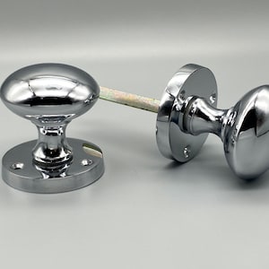 May include: A pair of polished chrome door knobs. Each knob features an oval design and a circular base with screw holes. The knobs are joined by a metal spindle. The hardware is reflective and appears unused.