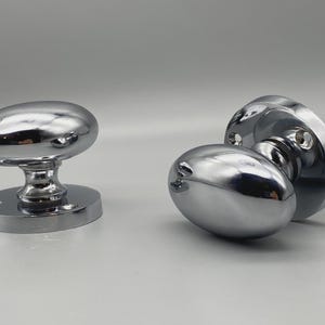 Puede incluir: Dos pomos de puerta cromados pulidos. Uno está en posición vertical, mostrando la forma ovalada y la base. El otro está unido a una placa y husillo, tumbado de lado. Los pomos tienen una superficie reflectante.
