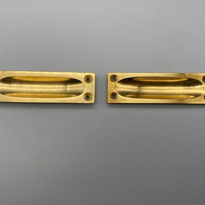Könnte beinhalten: Zwei rechteckige eingelassene Messinggriffe mit ovalen Fingermulden. Jeder Griff hat vier Schraubenlöcher. Die Griffe sind goldfarben und vor einem grauen Hintergrund platziert.