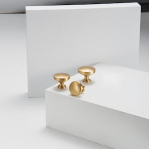 May include: Three gold-coloured cabinet knobs with a round top and a short, cylindrical base are displayed on a white surface. The knobs are arranged in a group, showcasing their design and finish. The background is a minimalist white.
