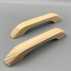 Könnte beinhalten: Zwei unfertige Holzschrankgriffe. Die Griffe sind hell gefärbt, mit einem geschwungenen Design und einer glatten Oberfläche. Sie sind aus Holz gefertigt und können gebeizt oder lackiert werden. Die Griffe sind etwa 15 cm lang.