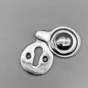 May include: A polished, silver-toned escutcheon and door pull hardware set. The escutcheon features a keyhole and two screw holes. The door pull is circular with a raised central element. The hardware is set against a grey backdrop.