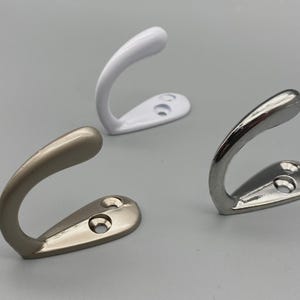 May include: Three metal coat hooks in white, silver, and muted gold finishes. Each hook has a curved design with a teardrop-shaped base and two screw holes. These hooks are designed for hanging coats, bags, or other items.