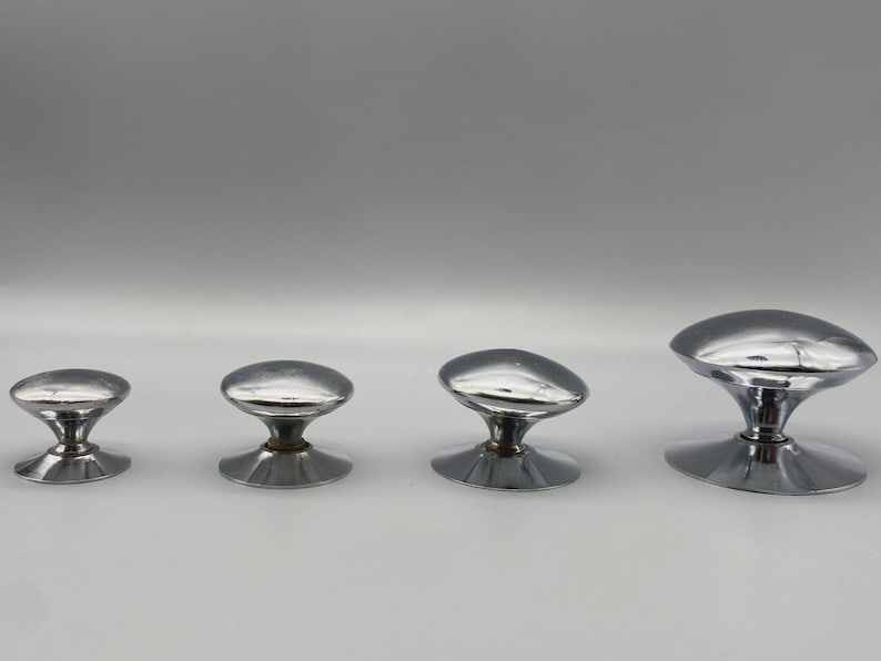 May include: Four chrome cabinet knobs of varying sizes are arranged in a row. Each knob has a rounded, oval-shaped top and a circular base. The knobs are reflective, with a shiny, metallic finish, and are set against a neutral background.