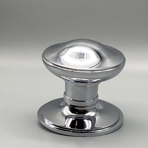 May include: A shiny, chrome-finished cabinet knob. The knob has a rounded top and a circular base, connected by a cylindrical stem. The polished surface reflects light, creating a bright, metallic appearance. This hardware is suitable for cabinets and drawers.