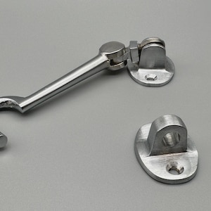 May include: A silver cabin hook and eye latch set. The set comprises a hook with a curved end, a pivoting bracket, and a separate eye plate. The hardware is designed for securing doors or windows.