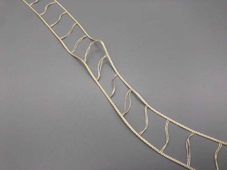 May include: A length of off-white decorative trim with a ladder-like design. The trim features a woven border with vertical and wavy strands connecting the edges. The background is a solid gray.